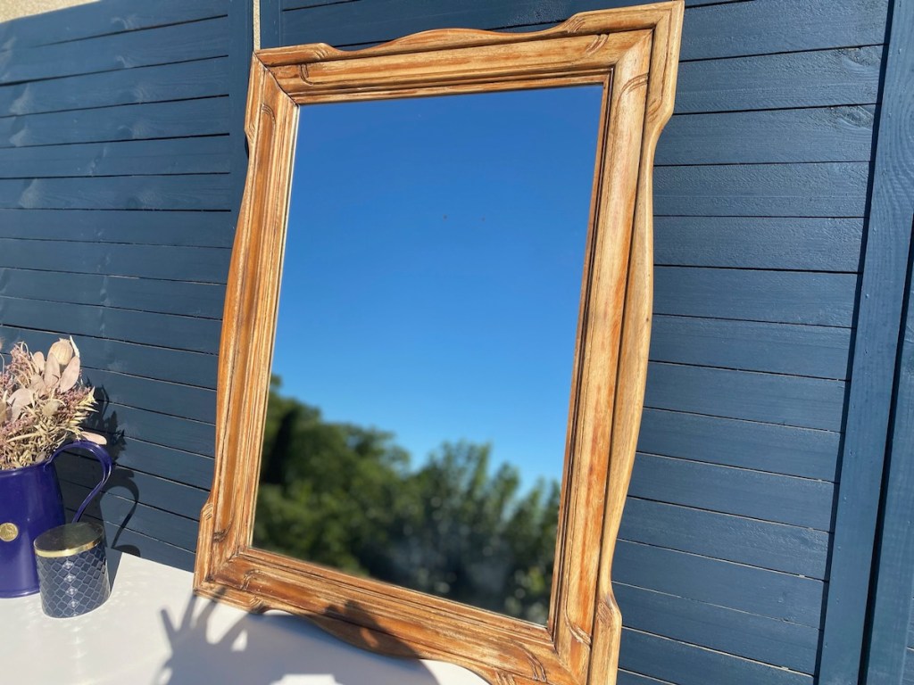 Un miroir élégant avec un cadre en bois sculpté, posé sur une table blanche, à côté d'un vase bleu avec des fleurs séchées. En arrière-plan, un mur bleu et un ciel dégagé.