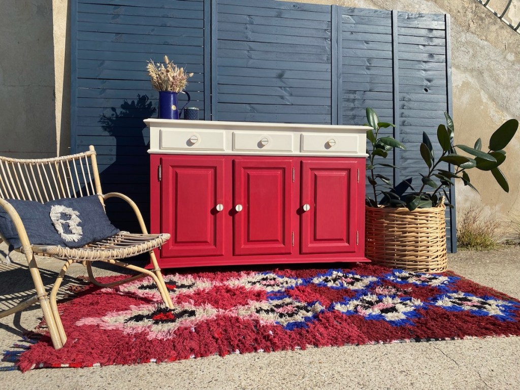 Un meuble rénové de couleur rouge avec des détails blancs, placé devant un mur de lattes bleu. Un fauteuil en rotin et un tapis coloré complètent la scène, avec une plante dans un pot en osier.