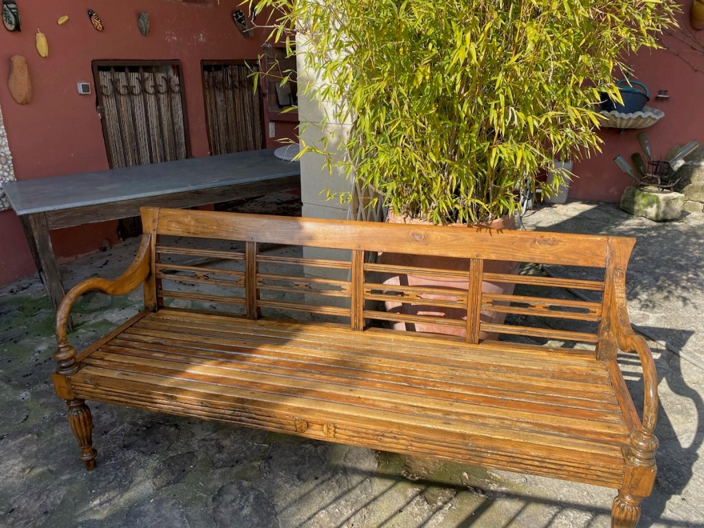 Un banc en bois ancien, avec des lattes apparentes et des accoudoirs, situé à l'extérieur avec un décor de murs en couleur chaude et des plantes alentour.