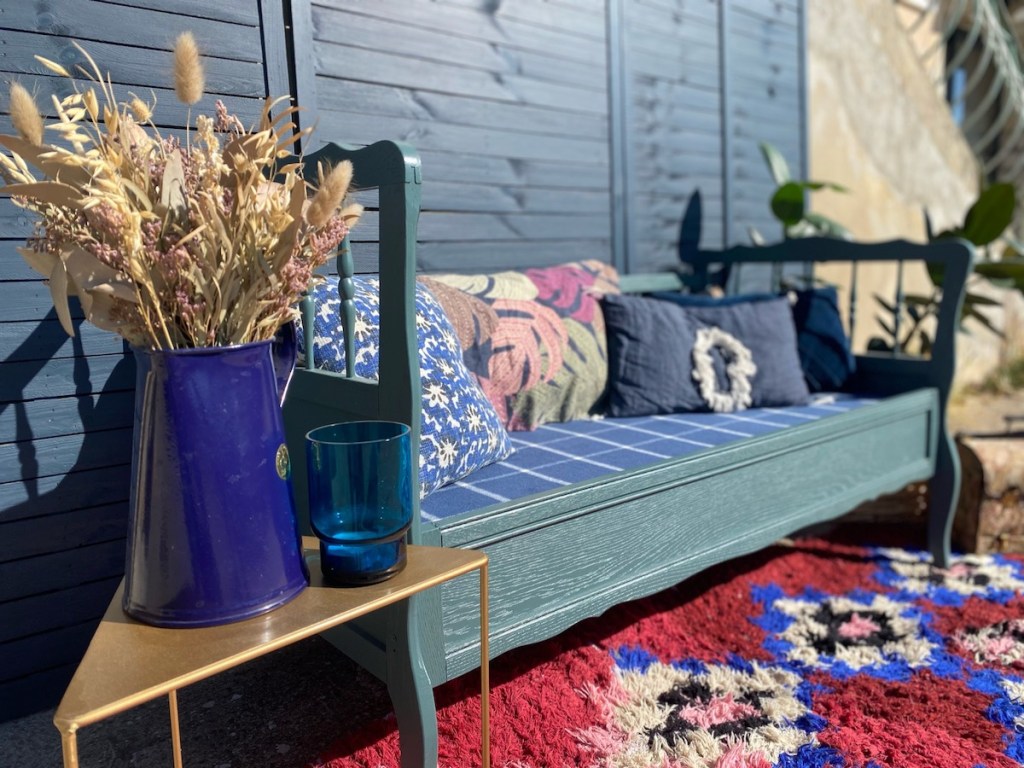 Un canapé coloré avec des coussins décoratifs, des fleurs séchées dans un vase bleu, et une petite table dorée. Le fond en bois donne une ambiance chaleureuse et accueillante.