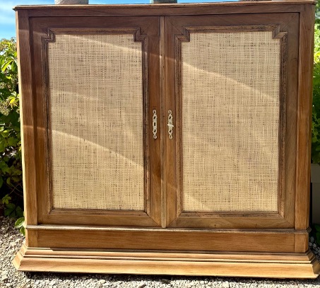 Ancien buffet en bois avec des portes en canage, teinté de beige, présenté à l'extérieur parmi des plantes.